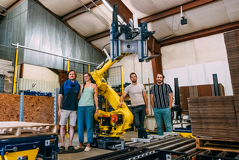 ein Foto von vier Personen, die neben einem Roboterarm zwischen Kartons aus Wellpappe stehen